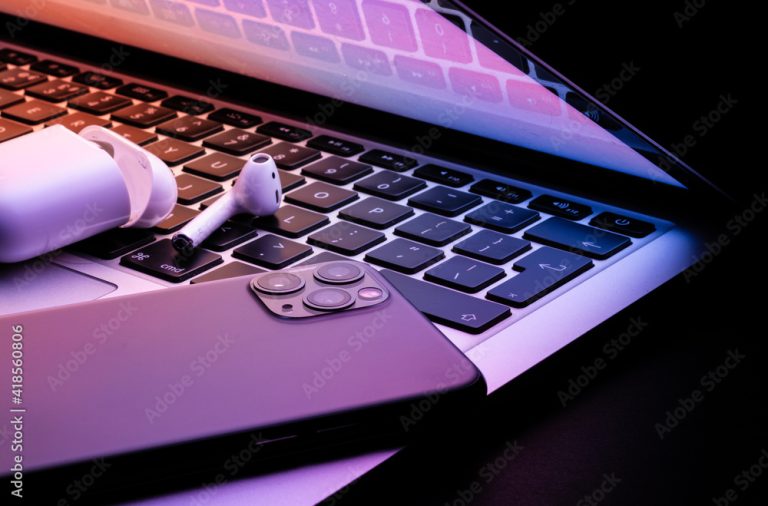 a laptop half closed in the dark with a phone and wireless earphones glowing